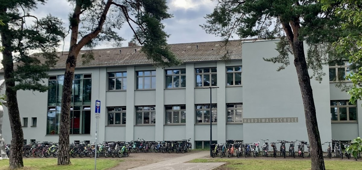 Außenansicht des Schulgebäudes der Hardtwaldschule. Das Gebäude hat einen grauen Farbton. Es ist zusehen, dass es ein Untergeschoss, Erdgeschoss, ein Obergeschoss mit Fenstern und ein Dachgeschoss ohne Fenster gibt. Vor dem Gebäude stehen viele Fahrräder und Bäume.   