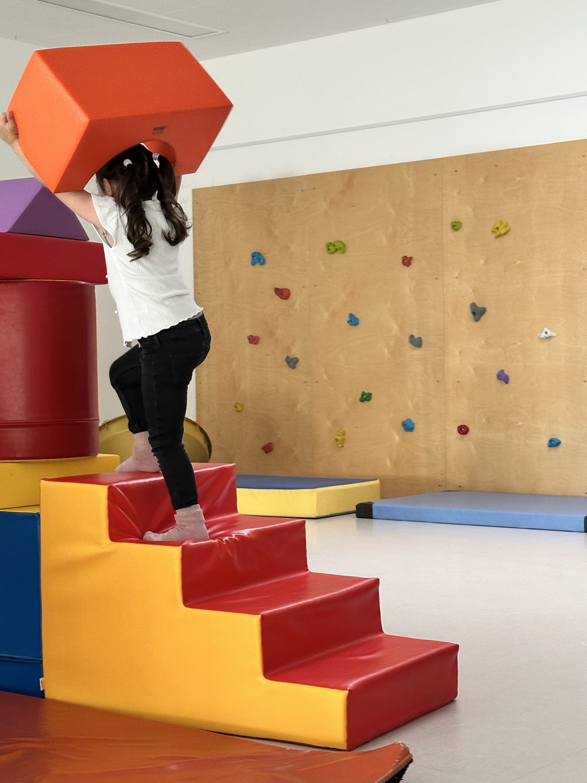 Kind steigt eine bunte Schaumstofftreppe hinauf und setzt einen großen, bunten Schaumstoffbaustein auf einen Turm aus anderen Bausteinen; im Hintergrund eine Bewegungslandschaft aus großformatigen, begehbaren bunten Schaumstoffelementen.