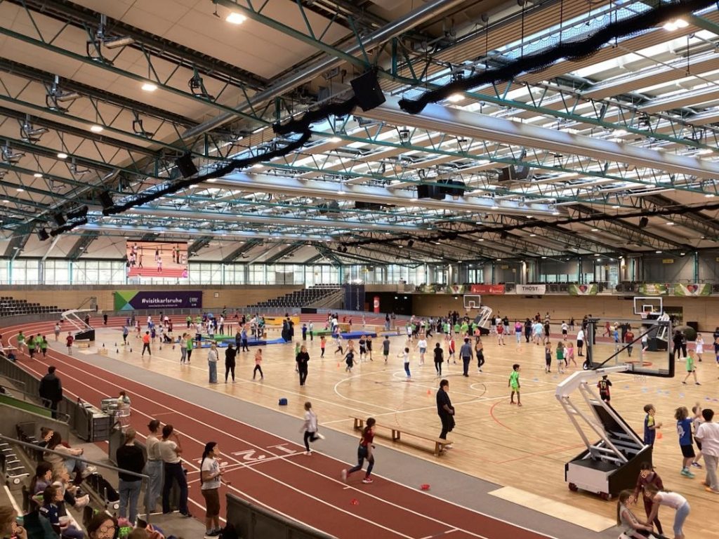 In einer großen Sporthalle, mit Aschenbahn und in inneren vielen Aufgebauten Sportstationen Bewegen sich viele Kinder und Erwachsene.