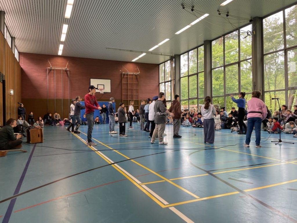 Tanzende Schüler und Schülerinnen, in einer Schulsportport Halle. Zuschauende stehen und sitzen an einer Wand und an einer großen Fensterwand.