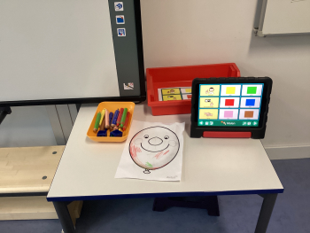 Arbeitsplatz mit Aufgabe zum Anmalen eines Luftballons; Tablet-PC als Kommunikationshilfe zeigt Farbauswahl, zur Auswahl und Benennung Farben.