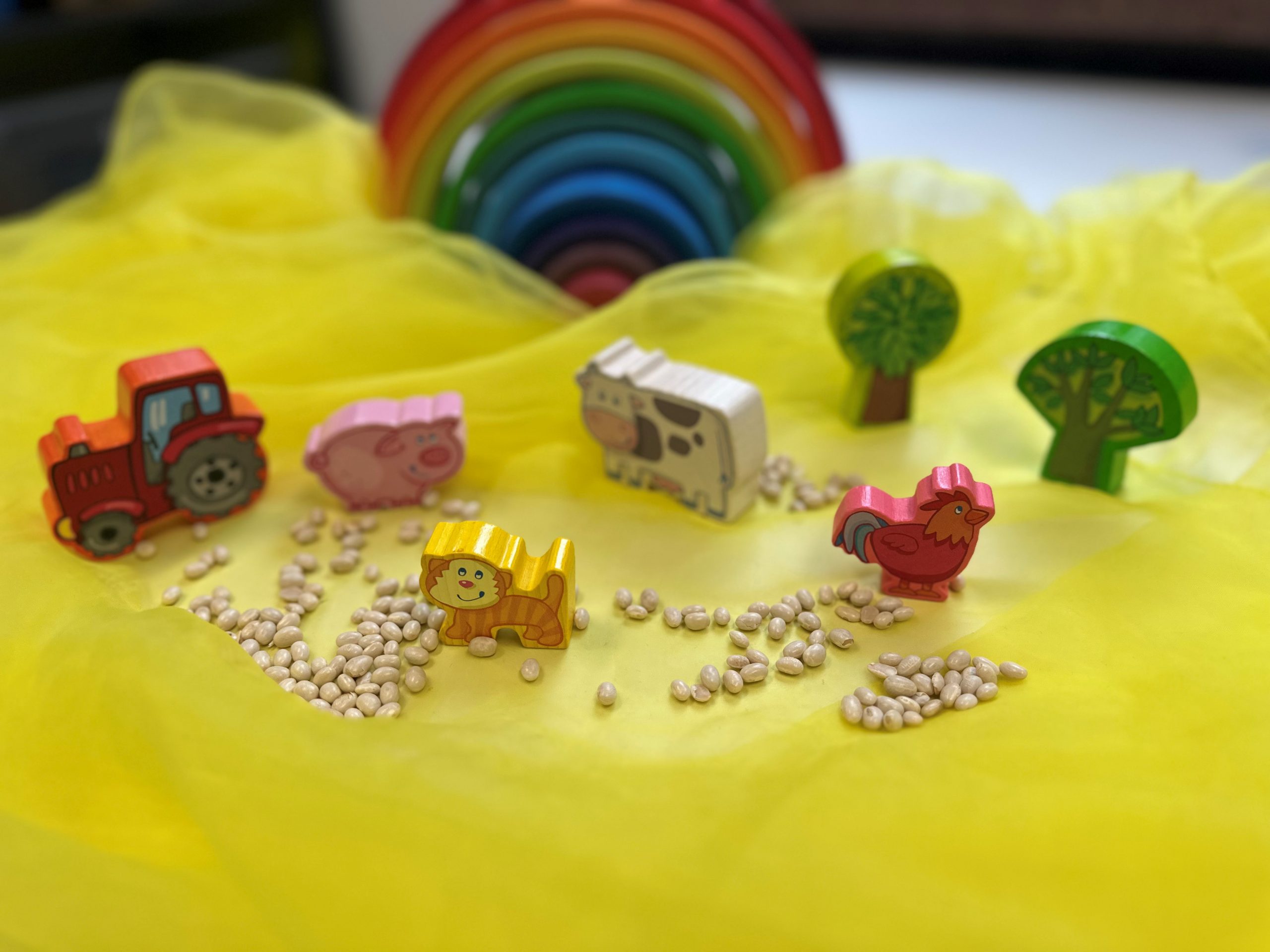 Holzspielfiguren (Kuh, Schwein, Schaf), ein kleiner roter Traktor, zwei Bäume und ein gestapelter Holzregenbogen stehen auf einem gelben Tuch.