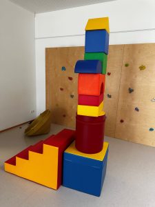 Bunte, große Schaumstoffbausteine aufgetürmt in einem Bewegungsraum; im Hintergrund eine Kletterwand.