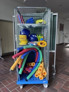 Gitterbox als Materialwagen gefüllt mit verschiedenen Schwimmhilfen und Schwimmutensilien (Bretter, Schwimmnudeln, Wasserbälle, Spielzeug).