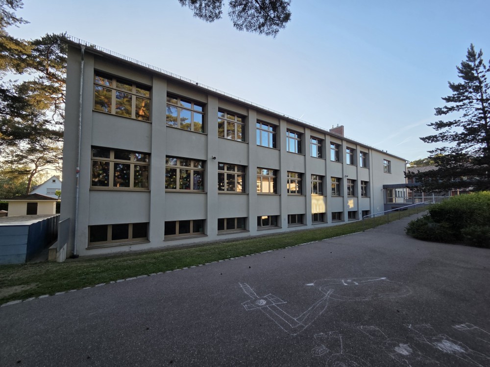 Schulgebäude der SBBZ Hardtwaldschule mit großer Fensterfront und Haupt­eingang, Ansicht vom Schulhof mit Gehweg ohne Treppen zum Haupteingang.