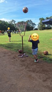Kinder und Jugendliche auf einem Sportplatz. Ein Kind wirft einen Basketball in einen Korbballständer.