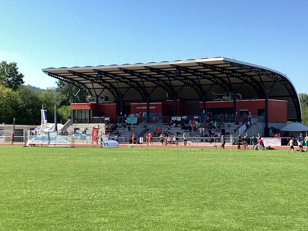 Blick auf den Sportplatz am Hans-Bretz-Stadion in Ettlingen: Rasenfeld mit Menschen, die an einer Sportveranstaltung teilnehmen, im Hintergrund das Stadion mit Zuschauern unter dem überdachten Bereich, viele Personen sitzen oder stehen.