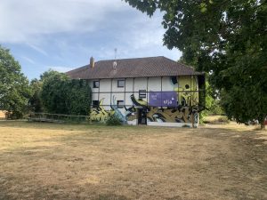 Außenansicht des Kinder- und Jugendhauses Trafohäus in Karlsruhe-Neureut: ein altes Fabrikgebäude, bunt besprüht, links mit grüner Bepflanzung und auf einer Wiese mit Bäumen.