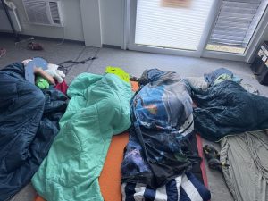 Vier schlafende Schülerinnen in Schlafsäcken auf Isomatten am Boden. Die Gesichter sind unkenntlich gemacht. Im Hintergrund sind heruntergelassene Rolläden zu sehen und eine Klimaanlage ist erkennbar.